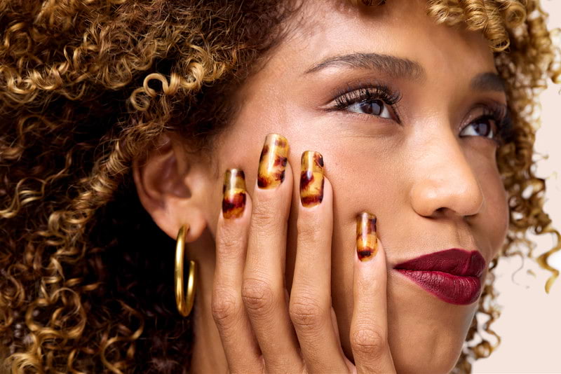 A woman with gold hoop earrings and curly hair resting a tortoiseshell manicure against her cheek.