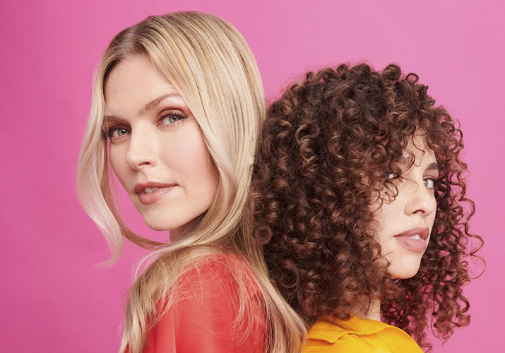 a woman with blonde straight hair standing back to back with a woman with warm brown curly hair 